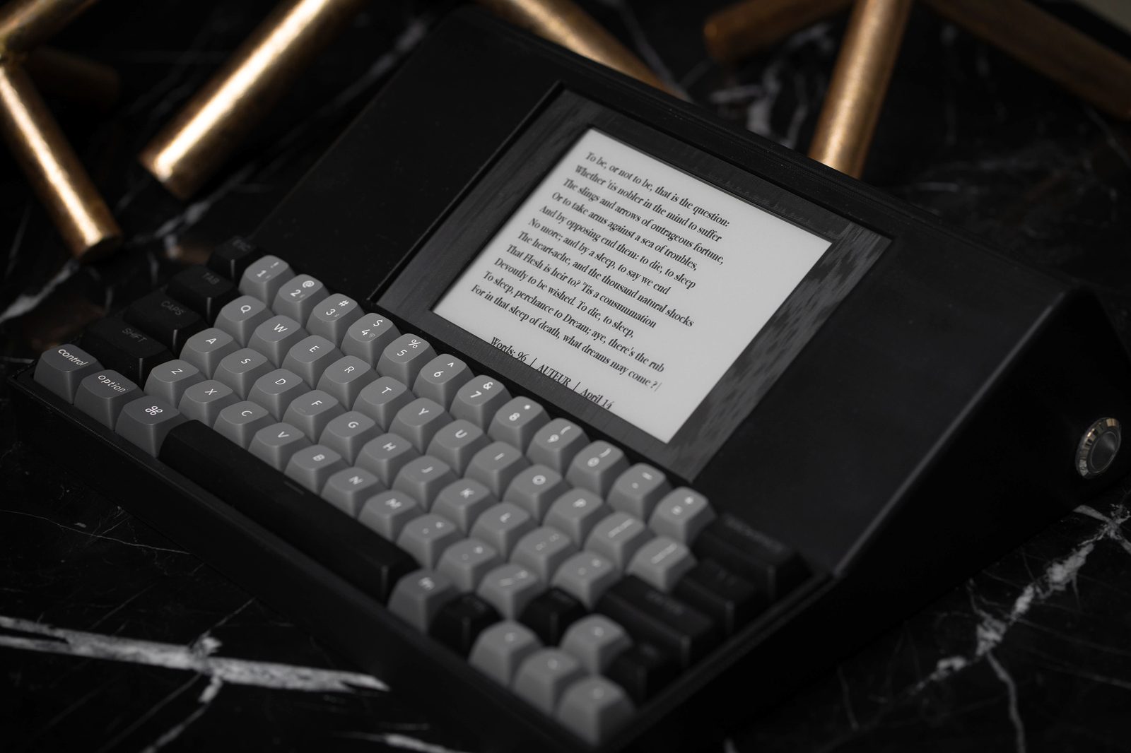 The AUTEUR prototype on a black marble surface, e-ink display showing the To be, or not to be soliloquy from Hamlet, with brass picture frames in the background.