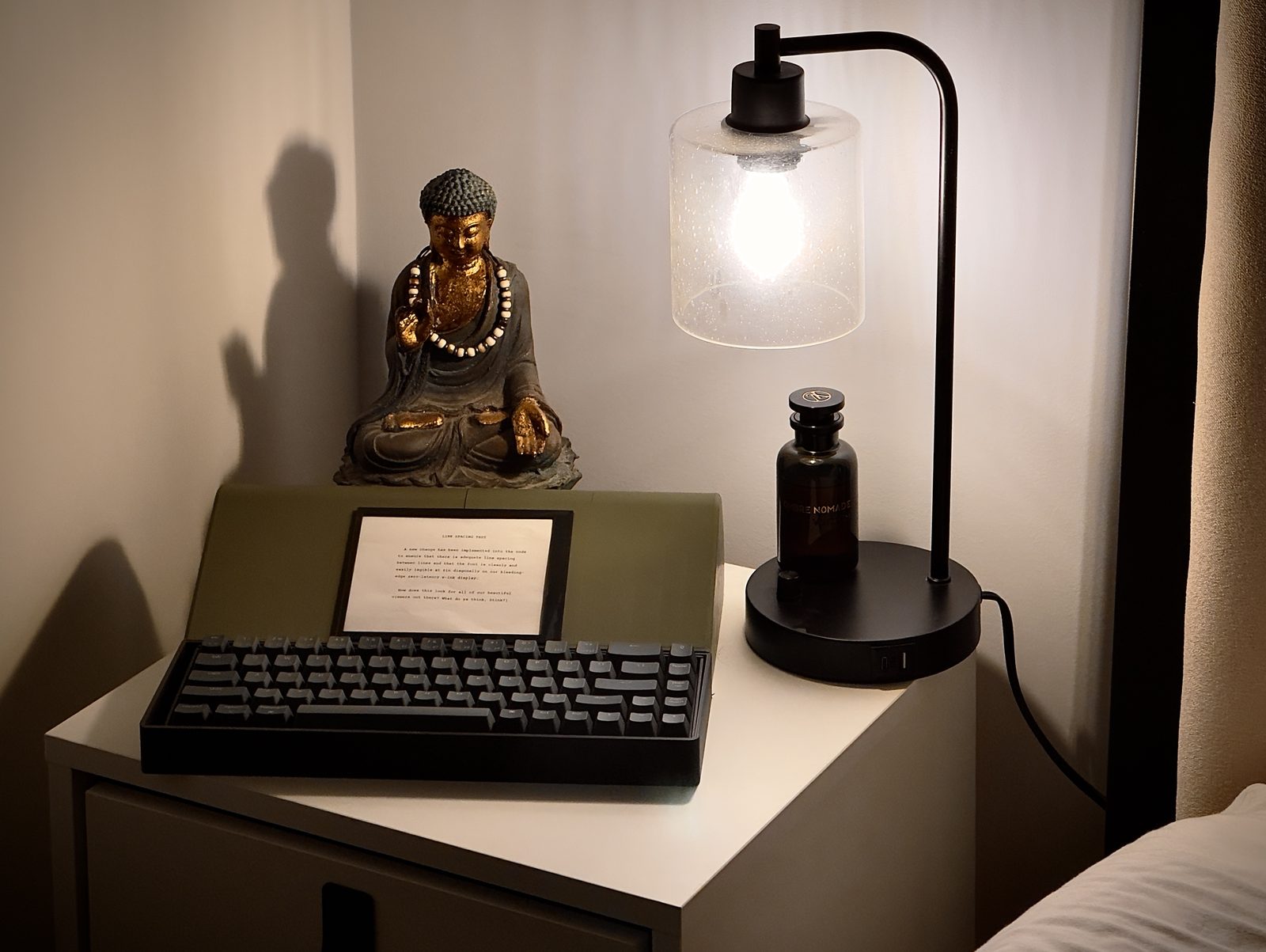 An early AUTEUR prototype on a bedside table, lit by a lamp, with a small Buddha statue beside it. A printed line-spacing test page is propped on the device.