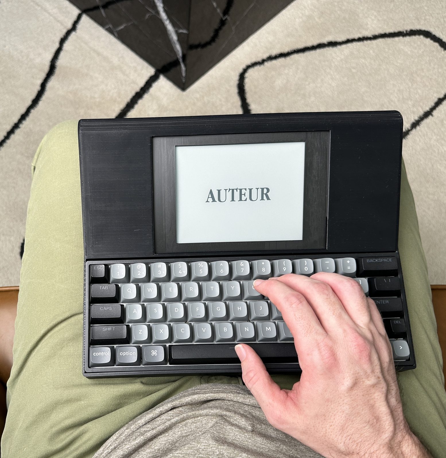 A hand resting on the AUTEUR keyboard, the device on the user's lap, the e-ink display showing the AUTEUR splash screen.